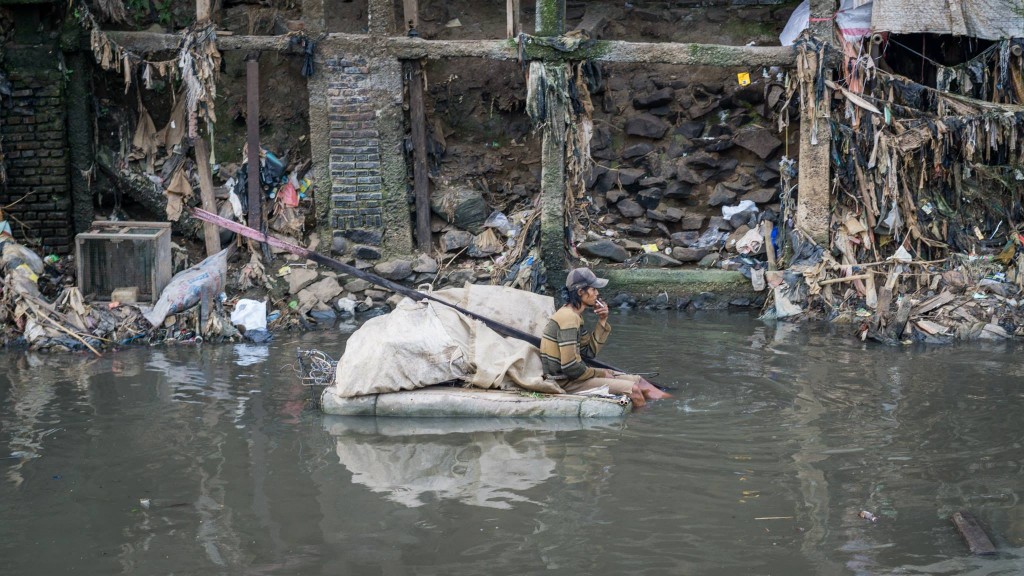 A river-based rubbish savager (pemulung) floating in the filthy river with his cigarette.